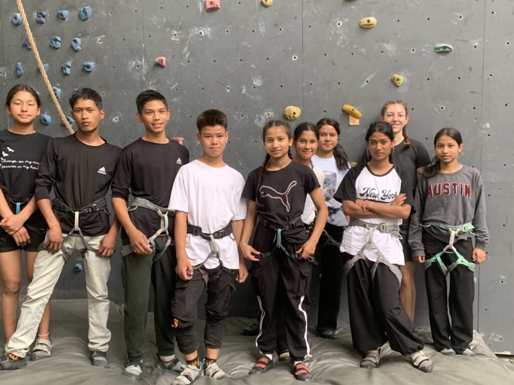 Bérénice stands with nine of the older Ama Ghar children in front of a rock climbing wall. All children are wearing harnesses and there are multiple colored climbing holds on the wall and a climbing rope hanging in the background.