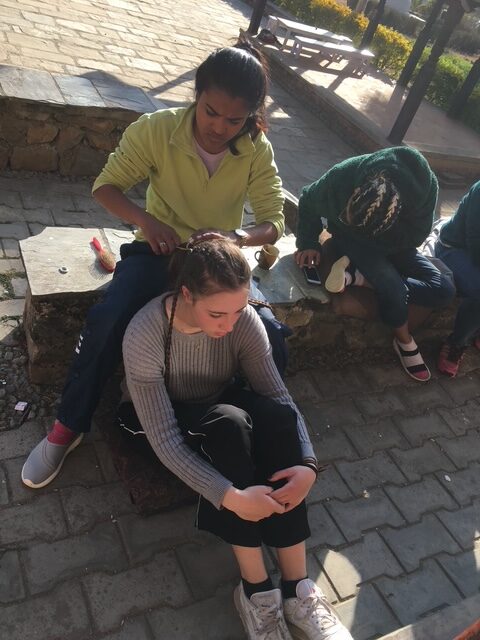 A teenage girl sits on the ground while another girl sits on a ledge behind her and braids her hair.