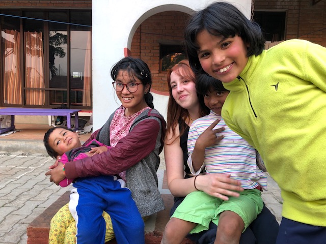 Two girls sit with a younger girl and boy on their laps. Another girl on the right leans into the frame.